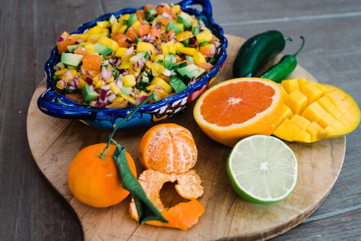 tigela azul de salsa de laranja, abacate e manga numa tábua de cortar com os ingredientes crus para a sua confecção