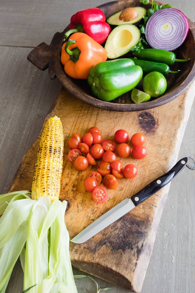 tigela de madeira de legumes numa tábua de cortar com uma espiga de milho torrado e alguns tomates cereja cortados pela metade