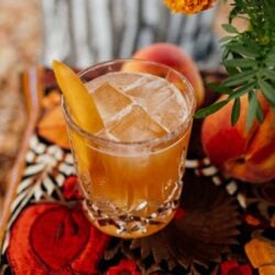 cocktail à moda antiga de pêssego bourbon num vidro cortado com pedras de cristal sobre uma paisagem de mesa vermelha e laranja com calêndulas
