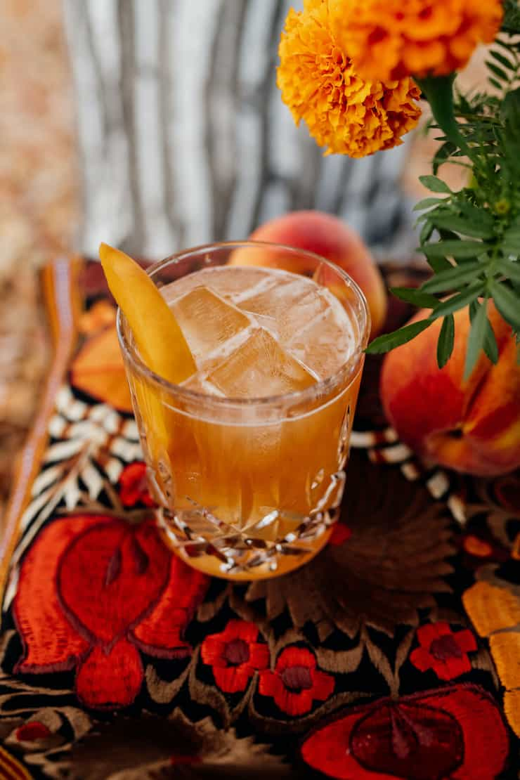 pêssego bourbon à moda antiga com guarnição de pêssego num copo de cristal sobre uma paisagem de mesa vermelha e laranja com calêndulas