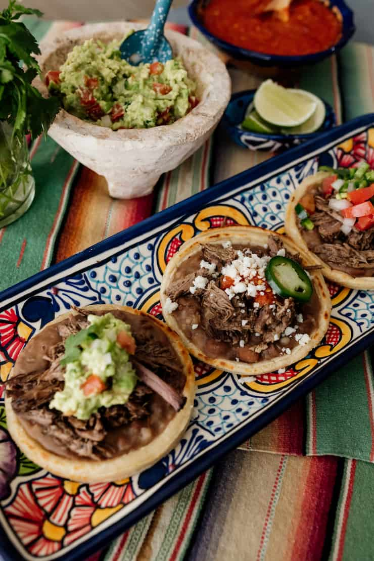 três maços de peito de vaca com cobertura mexicana numa travessa rectangular colorida com uma tigela de guacamole e salsa ao fundo