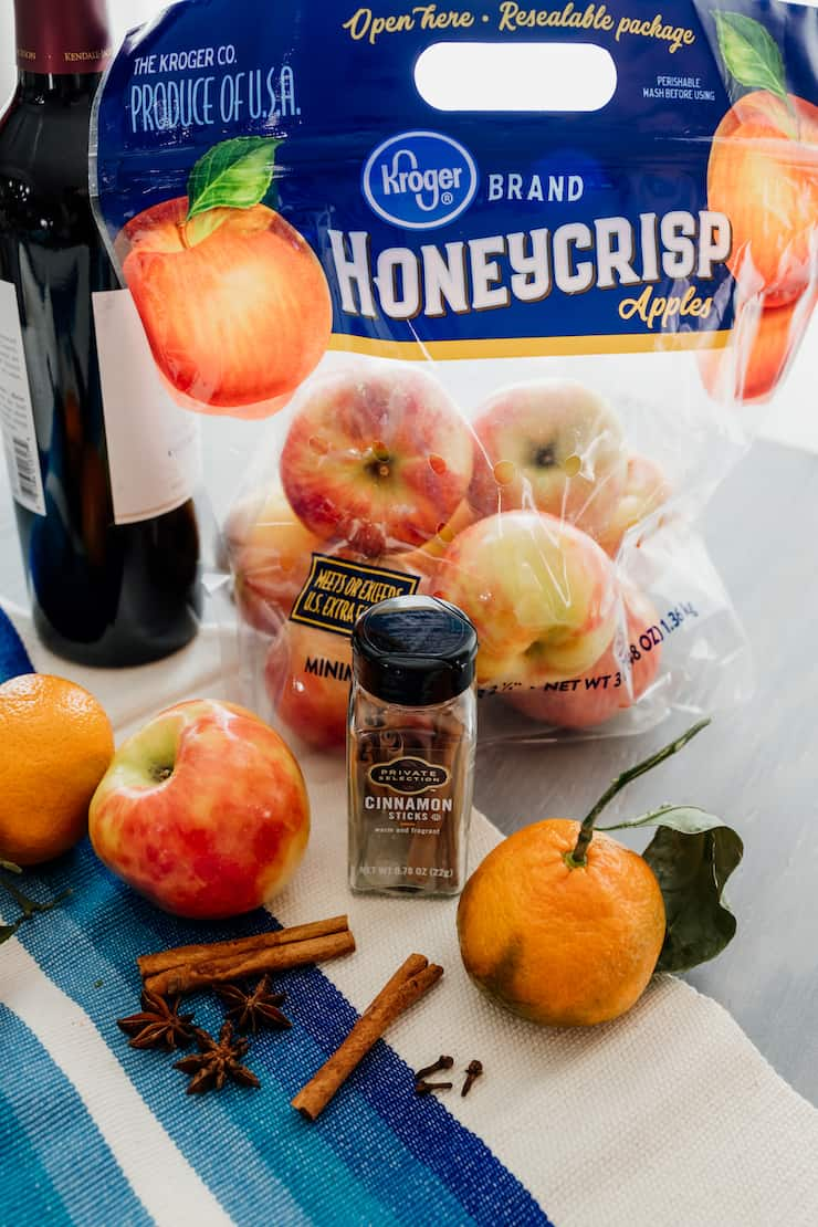 mise en place for mulled wine recipe - garrafa de vinho tinto, maçãs honeycrisp, pau de canela, anis inteiro e laranjas na videira