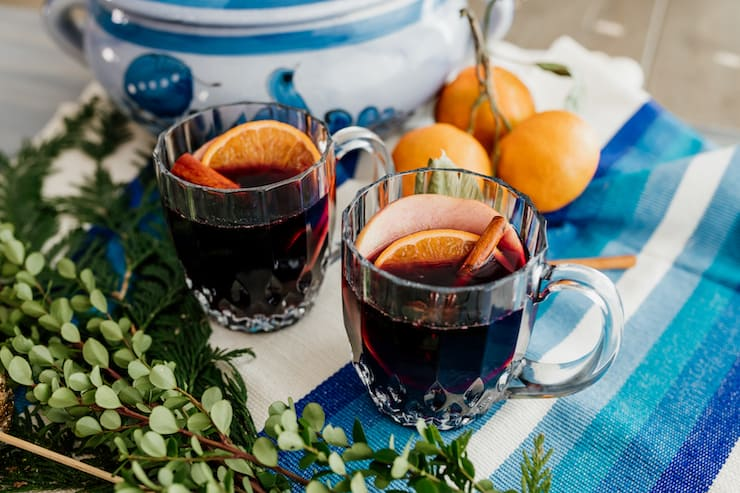 duas canecas de vinho temperado mulled com pau de canela e guarnições de fatias de frutas em uma paisagem de mesa azul e branca com verdura