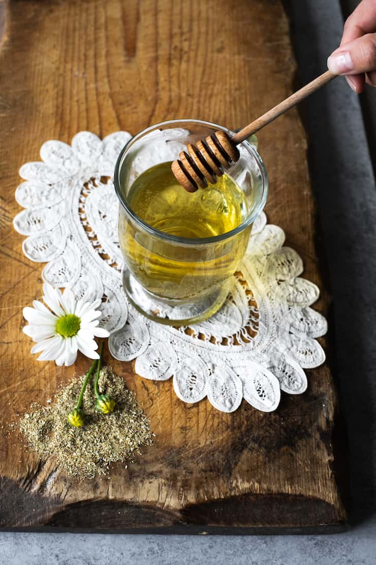 Té de manzanilla (chá de camomila) num copo de vidro transparente com um pauzinho de mel. O copo está em um doily vintage em uma superfície de madeira.