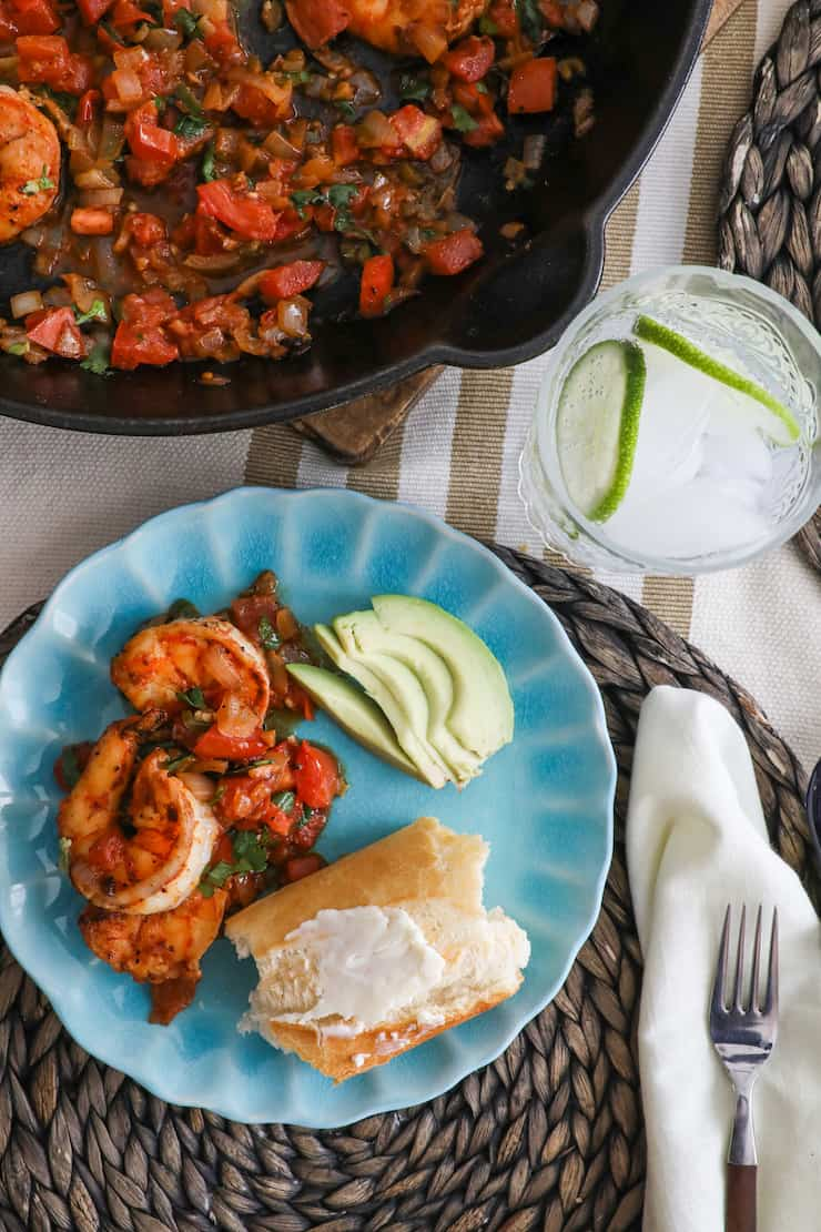 prato turquesa com camarões a la mexicana, abacate em fatias e pão crocante coberto com manteiga ao lado de uma frigideira cheia de camarões estilo ranchero mexicano