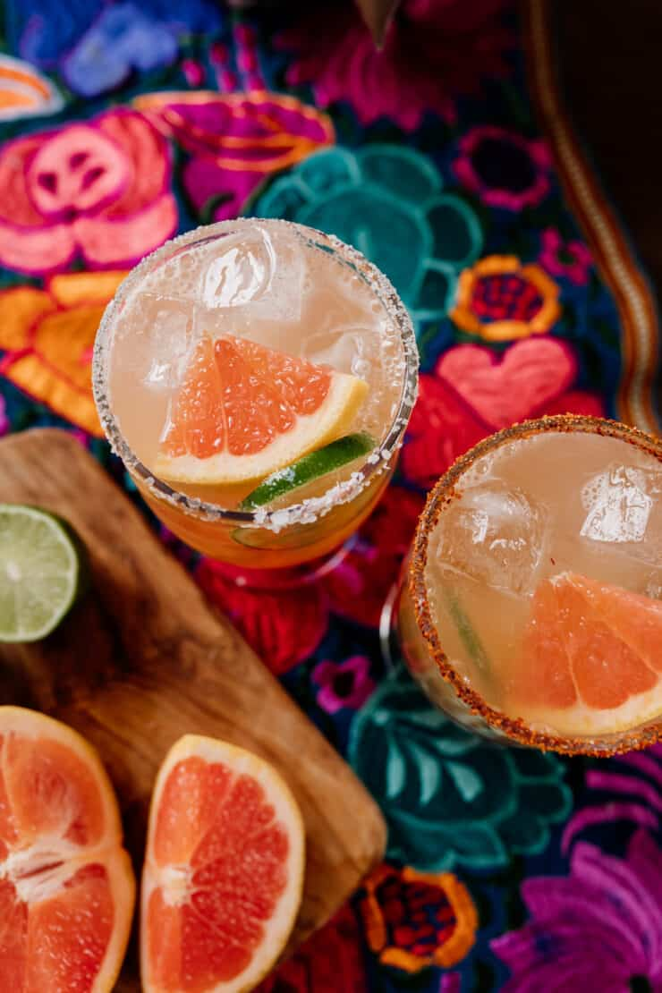 shot de cima para baixo de dois copos de cocktail preparados com Tajin e Sal com tequila reposado ao lado de cunhas de toranja fresca e lima num colorido tecido mexicano florido