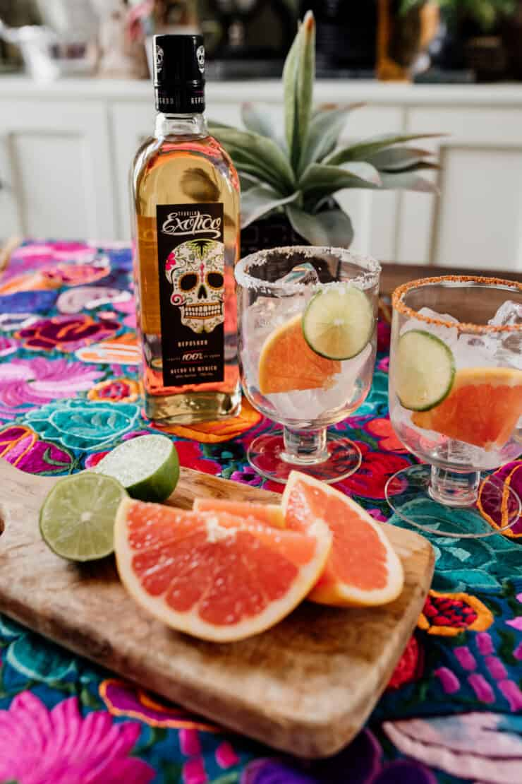 dois copos de coquetel preparados com Tajin e Sal com tequila reposado ao lado de cunhas de toranja fresca e lima em um colorido tecido mexicano florido