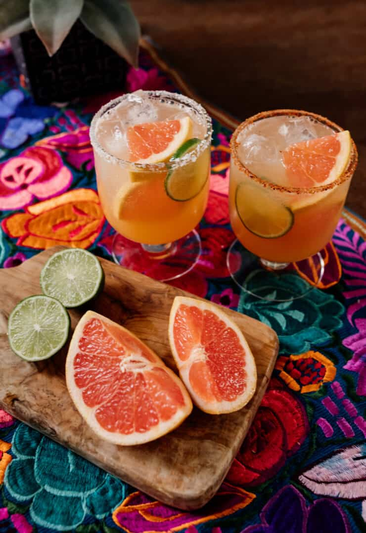 dois copos de cocktail cheios de palomas de reposado junto a cunhas de toranja fresca e lima num colorido tecido mexicano florido