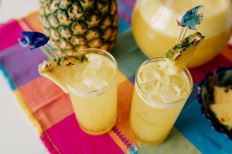 dois copos collins de agua fresca de ananás guarnecidos com agitadores de cocktail e cunhas de ananás fresco