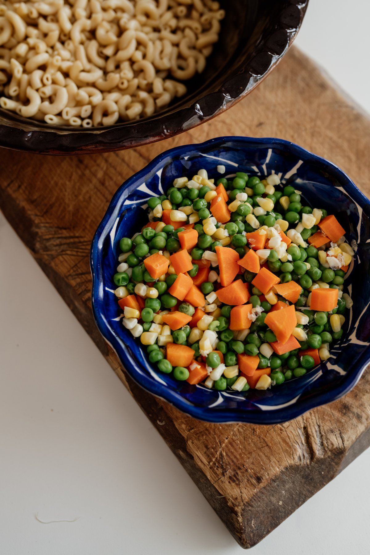 uma tigela azul mexicana cheia de cenouras, ervilhas e milho cozidos e macarrão cozido em uma tigela no fundo