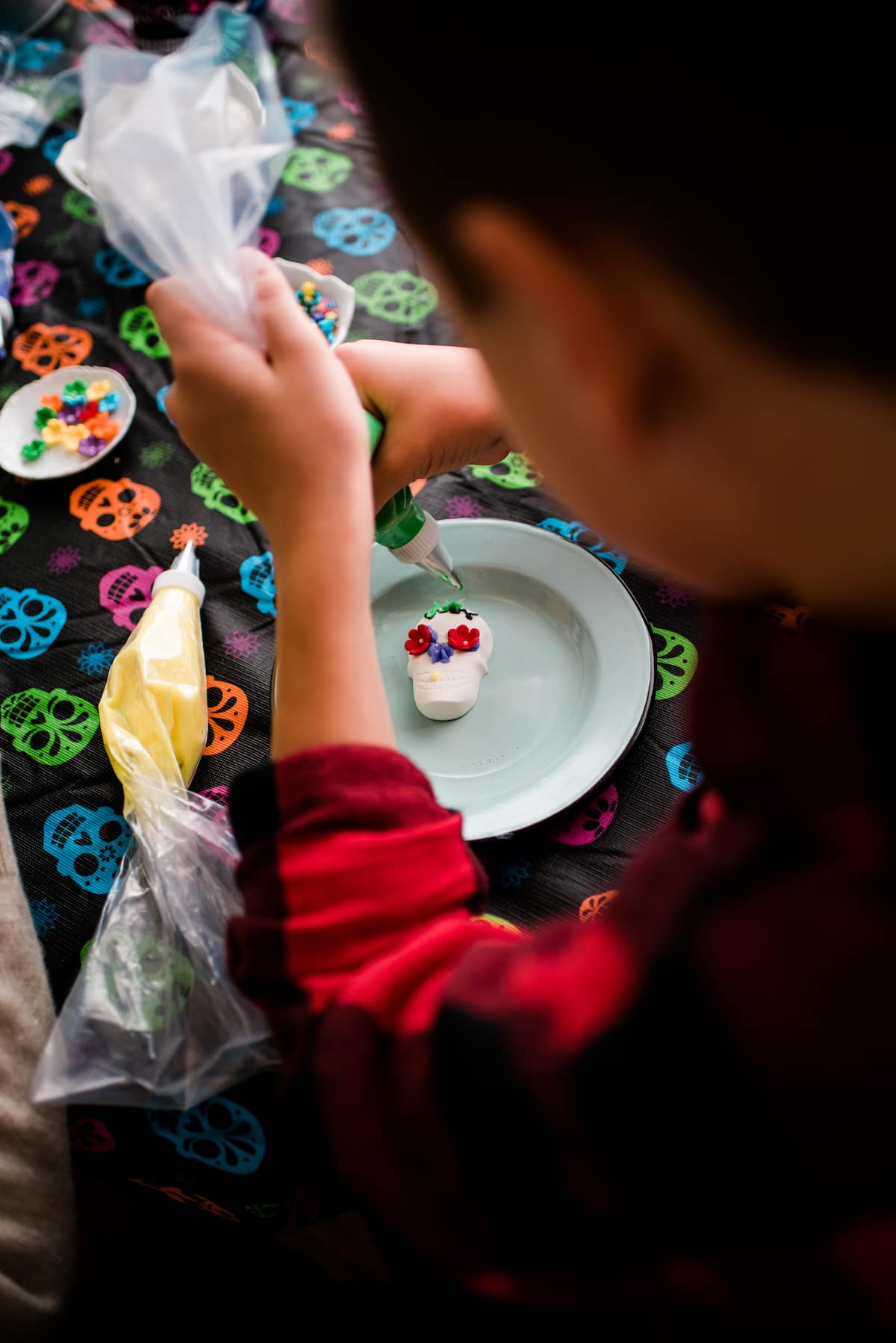 Uma foto aérea de um menino decorando seus crânios de doces Dia de los Muertos com cobertura colorida, flores de doces e salpicos em cima de uma toalha de mesa colorida no crânio.