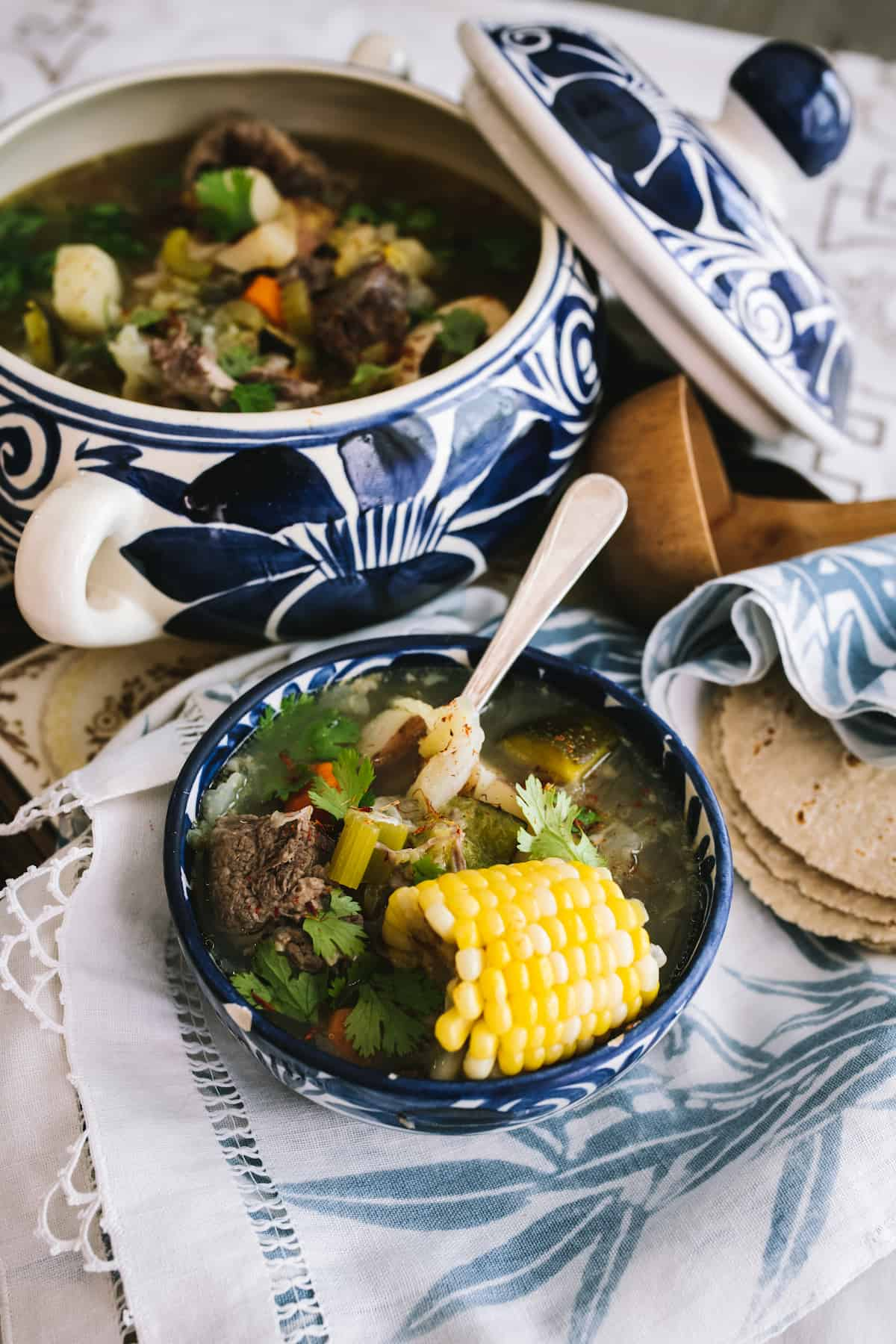 terrina mexicana azul e branca cheia de sopa de rabo de boi e uma pilha de tortilhas de milho e uma concha de madeira