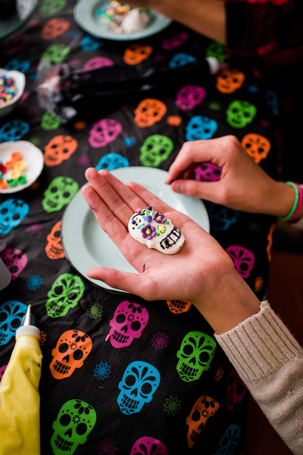 As crianças do autor decorando seus crânios de doces Dia de los Muertos com cobertura colorida, flores de doces e salpicos em cima de uma toalha de mesa do crânio