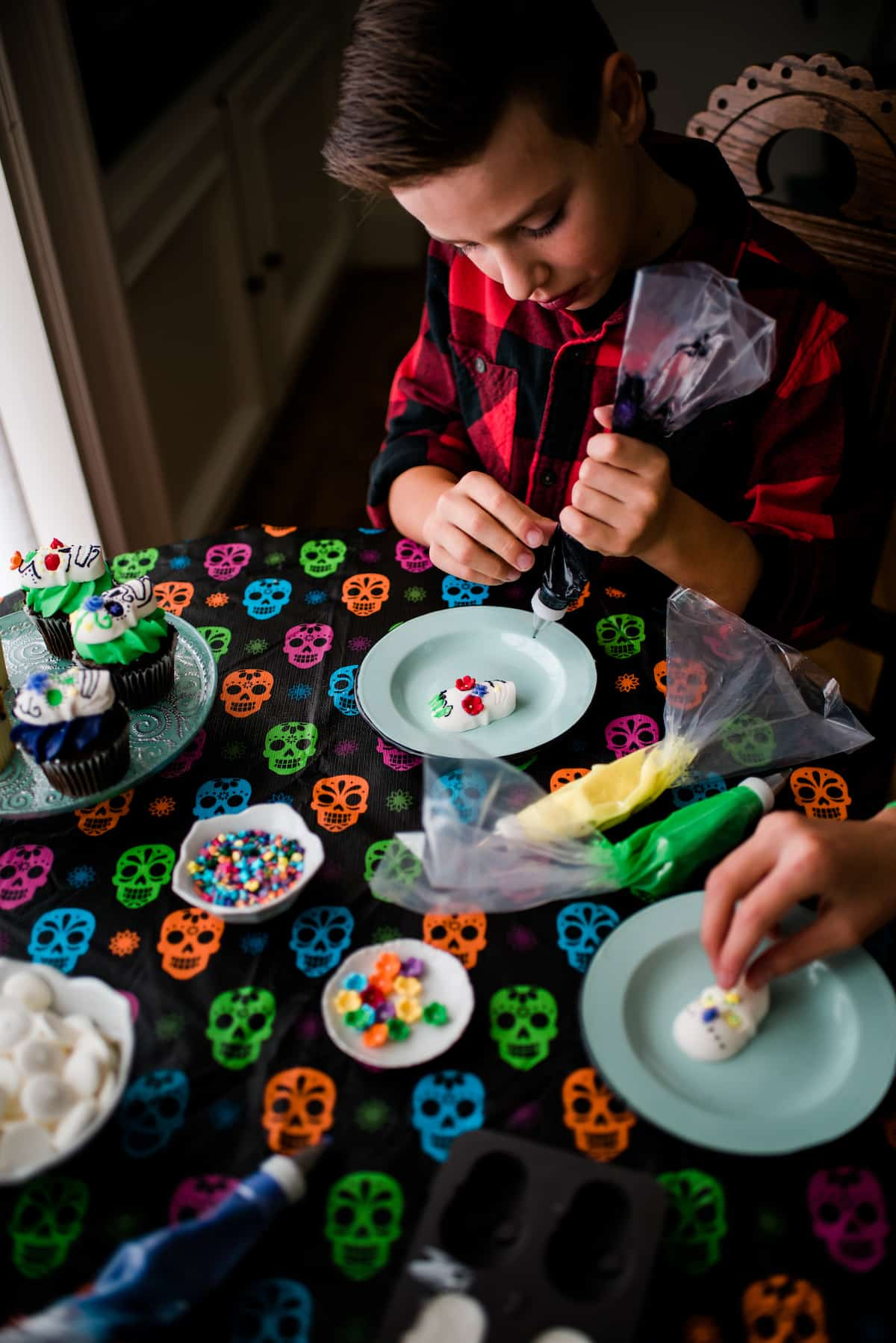 As crianças do autor decorando seus crânios de doces Dia de los Muertos com cobertura colorida, flores de doces e salpicos em cima de uma toalha de mesa colorida no crânio.