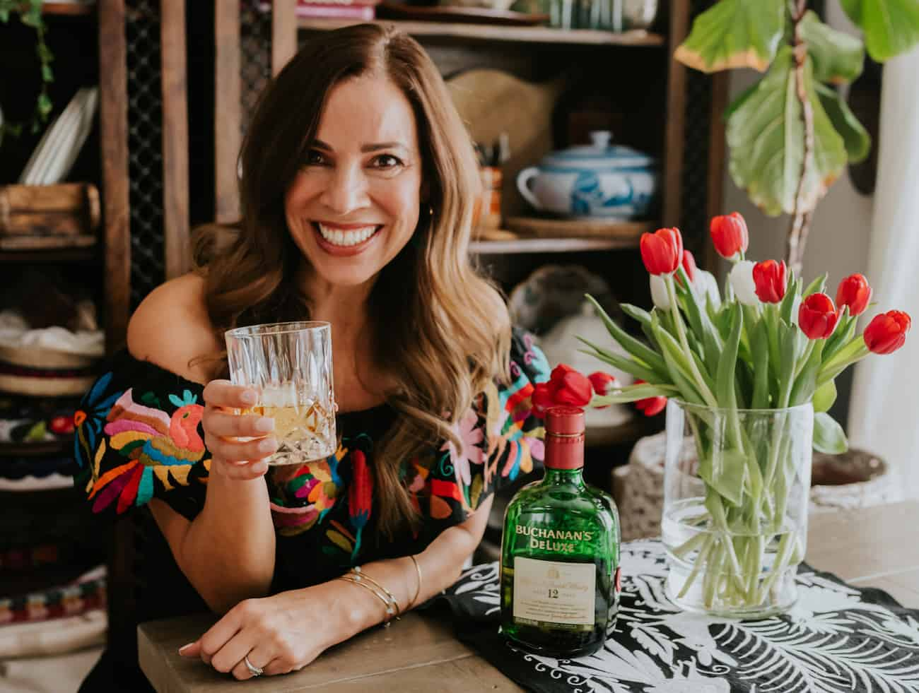 Yvette Marquez Latina Colorado blogger de comida segurando um copo de whisky com uma garrafa de Buchanan e um vaso de tulipas na lateral.