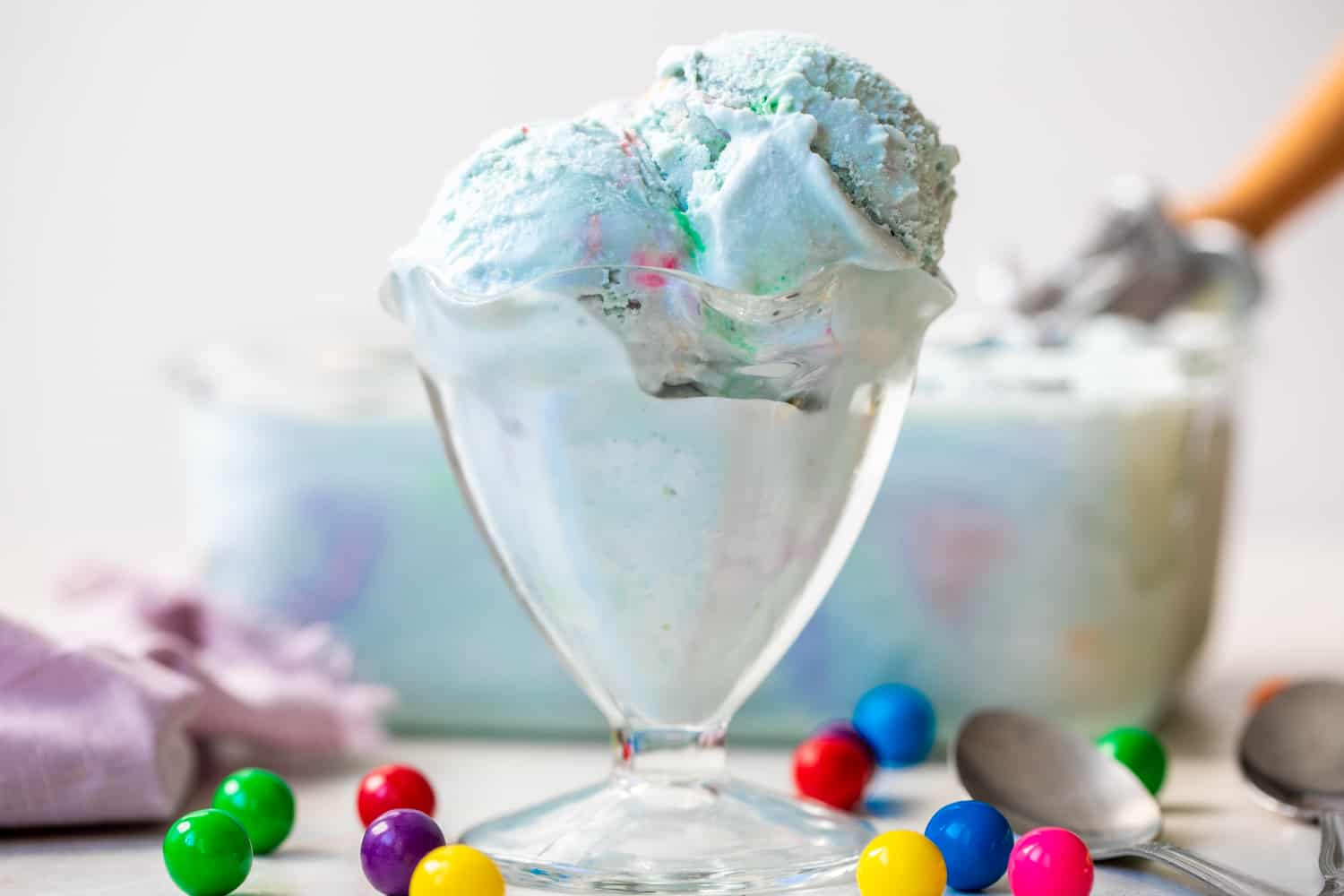 Copo para gelado de vidro cheio de gelado de pastilha elástica azul claro, recipiente de gelado no fundo e bolas de goma extra à volta do copo para gelado.