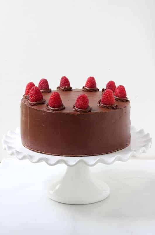 Bolo de Chocolate com framboesas frescas em cima sentado numa travessa de bolo branco.