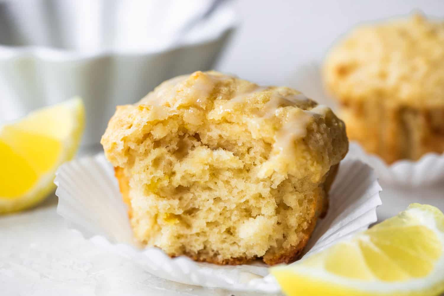 Imagem horizontal de muffin de limão assado com uma dentada faltando mostrando textura fofa. Cunhas de limão na lateral e uma tigela de gelo atrás do muffin.
