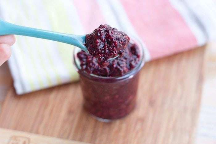 Compota mista de frutos silvestres Chia Seed Jam servido num pequeno frasco de vidro transparente com uma colher de porção azul clara na tábua de cortar madeira.