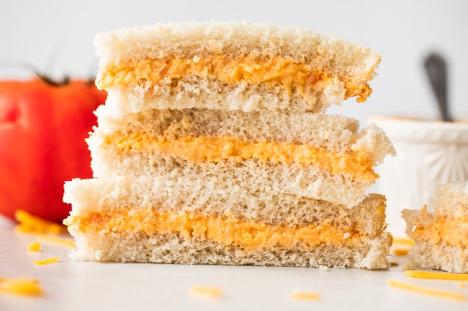 Pilha de sanduíches de tomate twiddle sobre pão branco, com queijo ralado no balcão ao seu redor, um tomate ao fundo e um ramequim recheado com twiddle de tomate.