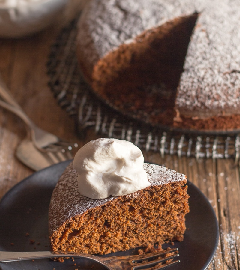 Bolo de bolo de gengibre numa placa de arame preto com uma fatia numa placa preta e um bocado de chantilly.