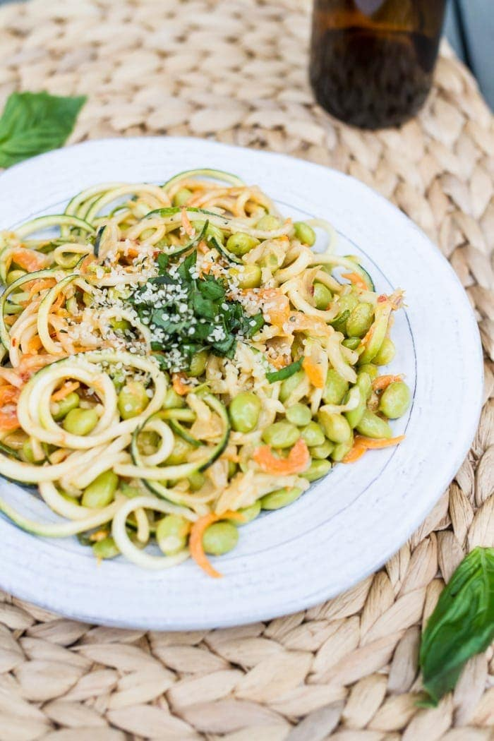 almofada de massa de abobrinha tailandesa, com edamame, coberta com folhas de manjericão frescas picadas, sobre um prato branco. Uma garrafa castanha está sentada no fundo.