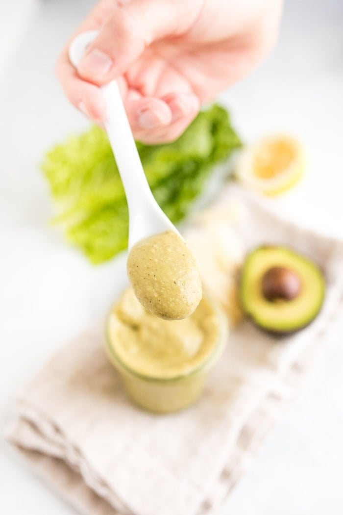 Mão segurando uma colher cheia de molho de abacate caesar acima de um frasco com o molho sobre uma mesa com alface e um abacate.