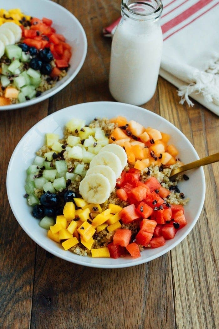 Taças de café da manhã de Canela Quinoa com frutas frescas picadas, servidas em tigelas brancas sobre mesa de madeira e copo de leite.