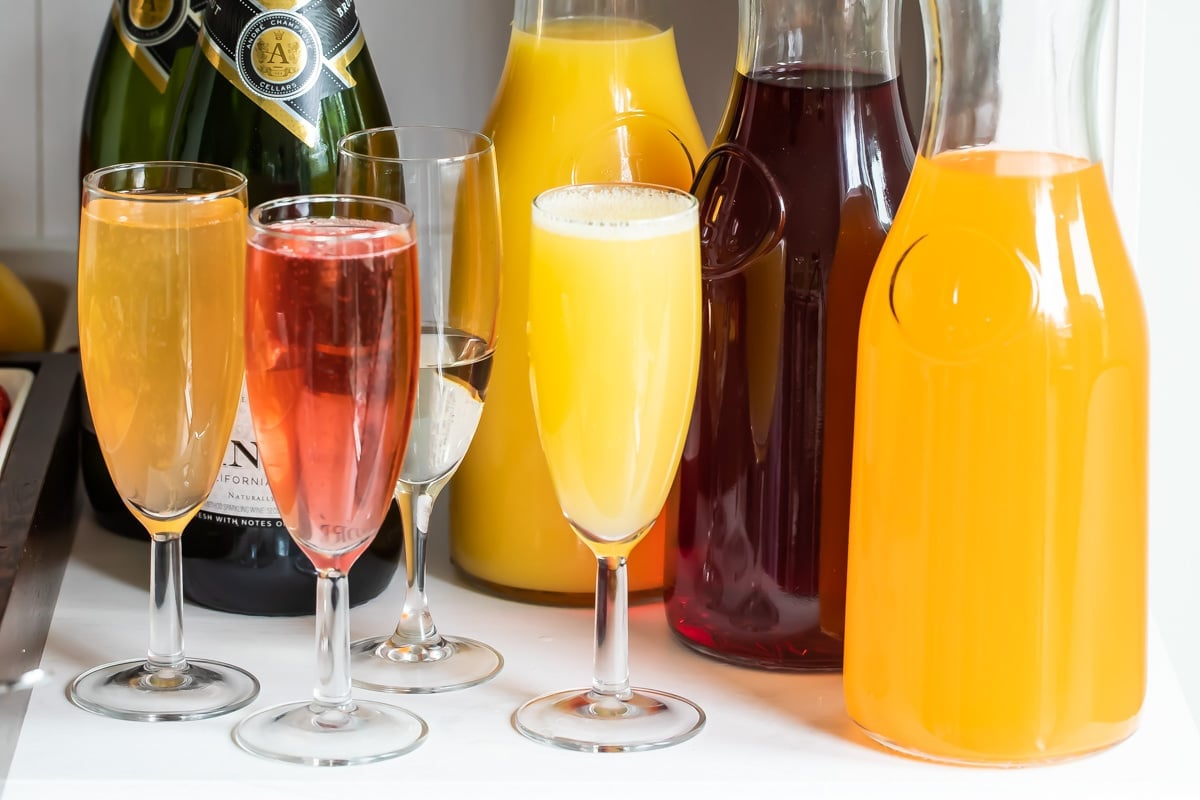 Três jarras com vários sucos e 4 flautas de champanhe com mimosas.