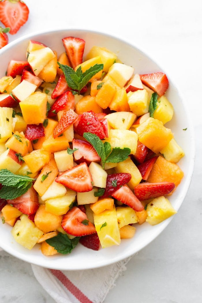 Tigela branca com salada de frutas incluindo morangos, abacaxi, melão, maçã e menta.