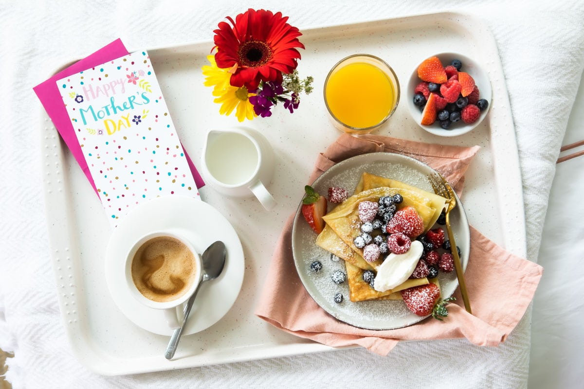Uma bandeja de café da manhã do Dia das Mães com crepes, suco de laranja, café, flores frescas, e um cartão de felicitações.