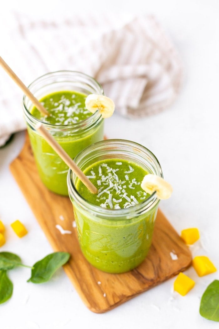 Dois potes de pedreiro Ball cheios com um batido verde sentado numa tábua de madeira com espinafres, mangas e pedaços de coco espalhados por todo o lado.