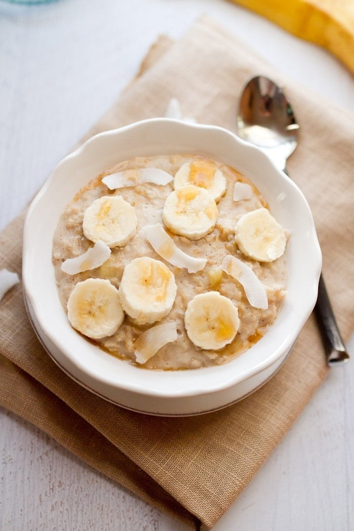 Uma tigela branca de farinha de aveia coberta com fatias de banana, pedaços de coco seco e uma garoa de mel. Uma colher está sentada ao lado da tigela.