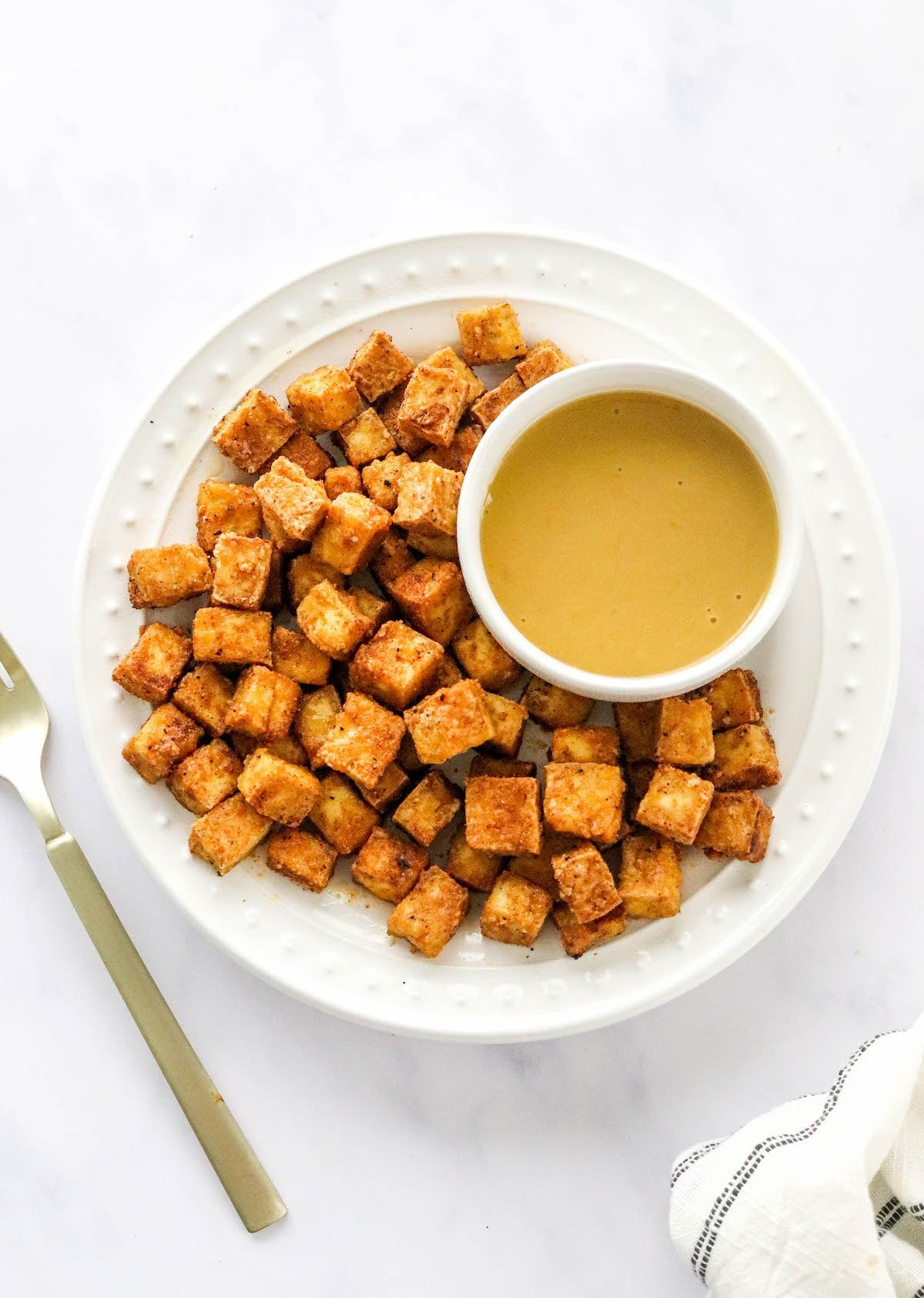 Uma foto aérea de um prato de tofu frito ao ar com um ramekin de molho de imersão. Um garfo dourado está ao lado do prato.