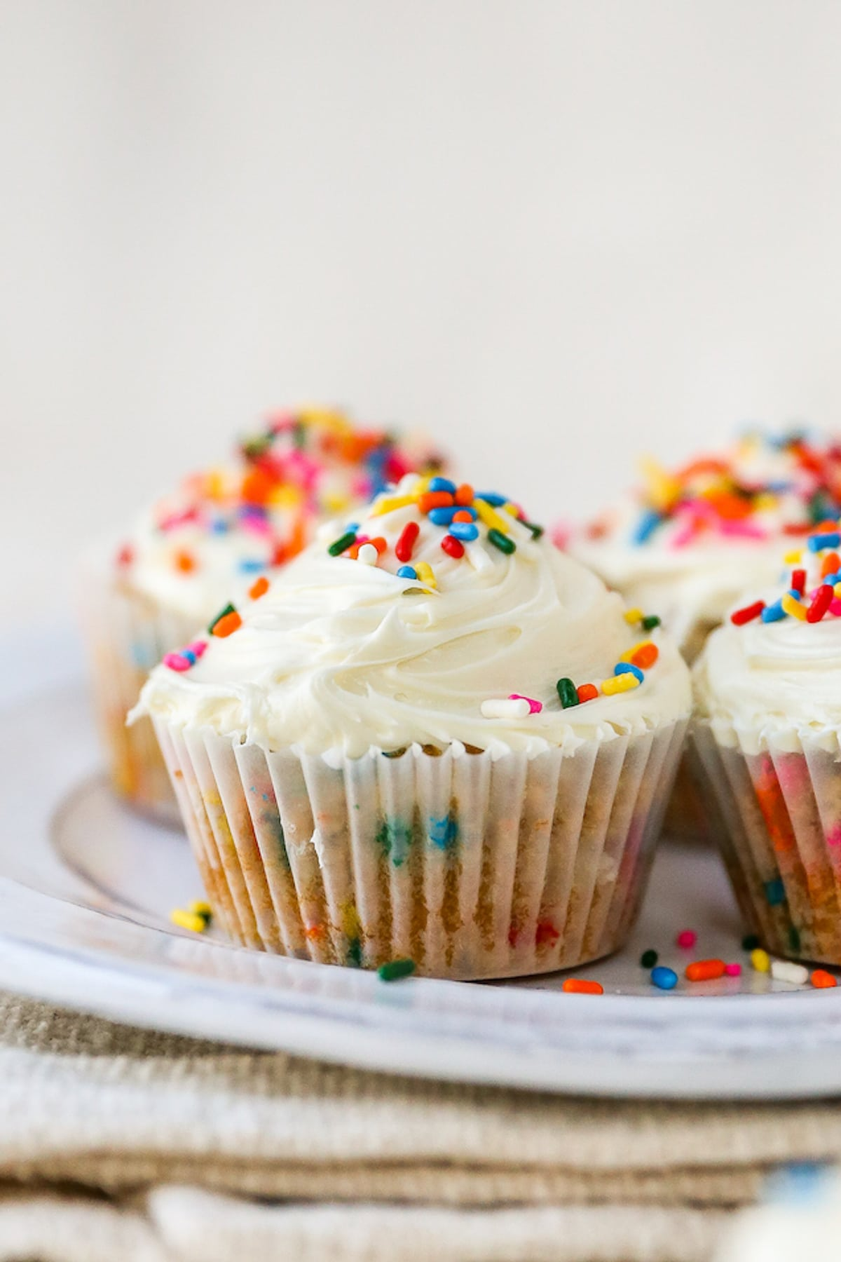 Quatro cupcakes de farinha de amêndoa embrulhados em forros de papel em um prato. Os cupcakes foram cobertos com cobertura de baunilha e polvilhos de arco-íris.