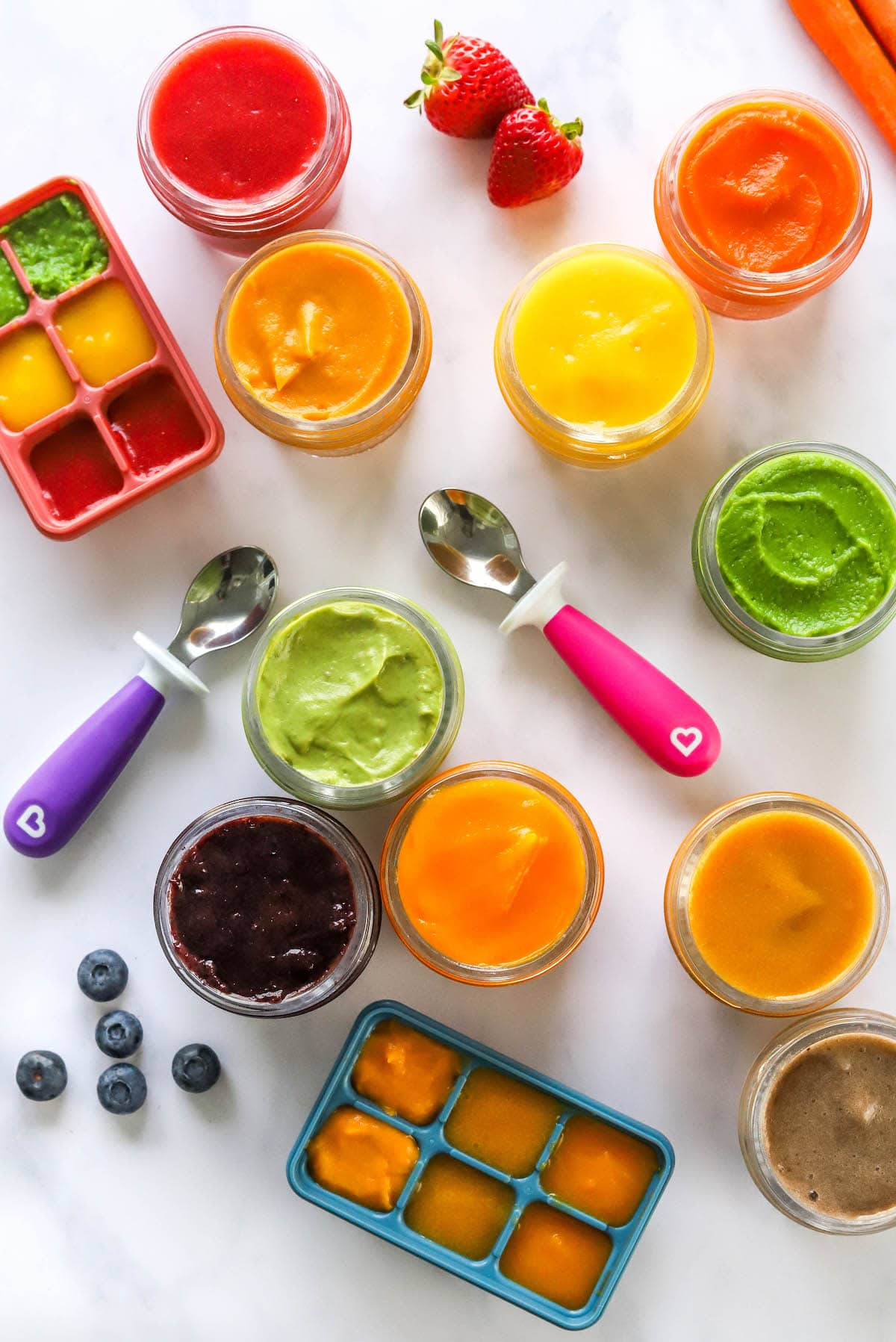 Dez variedades de purés de comida para bebé em frascos com morangos, mirtilos, colheres e tabuleiros de cubos de gelo espalhados à sua volta.