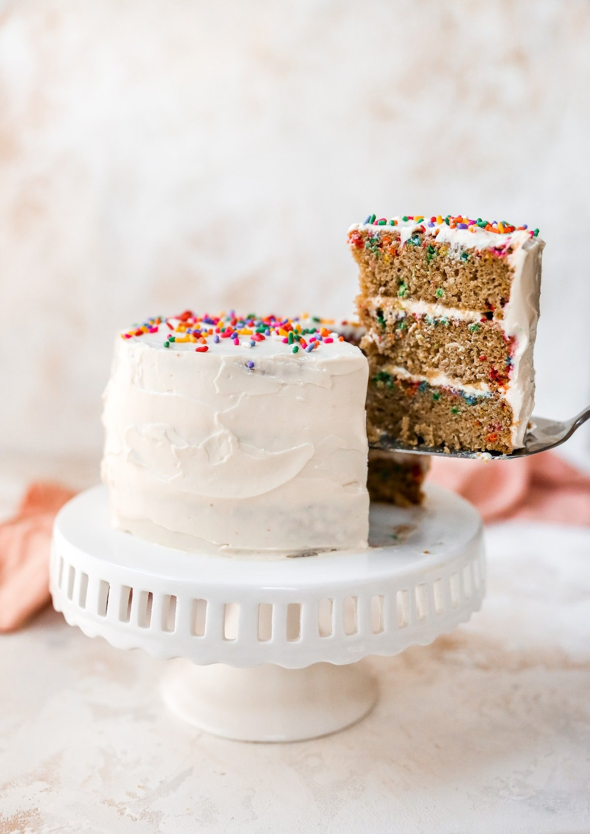 Um bolo sentado numa banca de bolos. O bolo está gelado em cobertura branca e coberto com salpicos de arco-íris. Uma fatia foi cortada e está sendo retirada, expondo as 3 camadas de bolo de funfetti com cobertura de cream cheese entre as camadas.