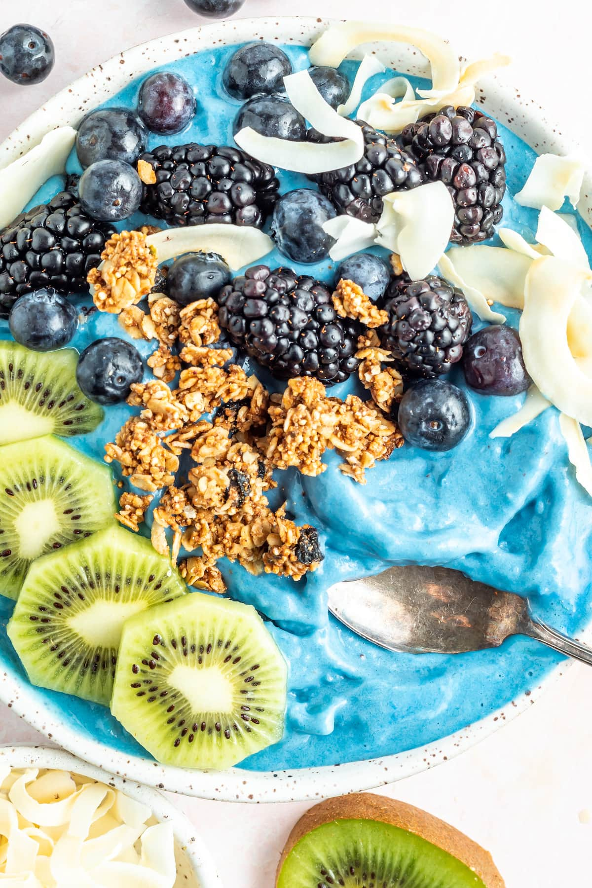 Foto aérea de uma tigela de smoothie majik azul coberta com kiwi, granola, amoras, mirtilos e flocos de coco.