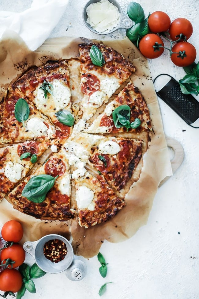 Pizza de couve-flor sobre papel pergaminho, colocada junto a um ralador de queijo e tomates frescos.