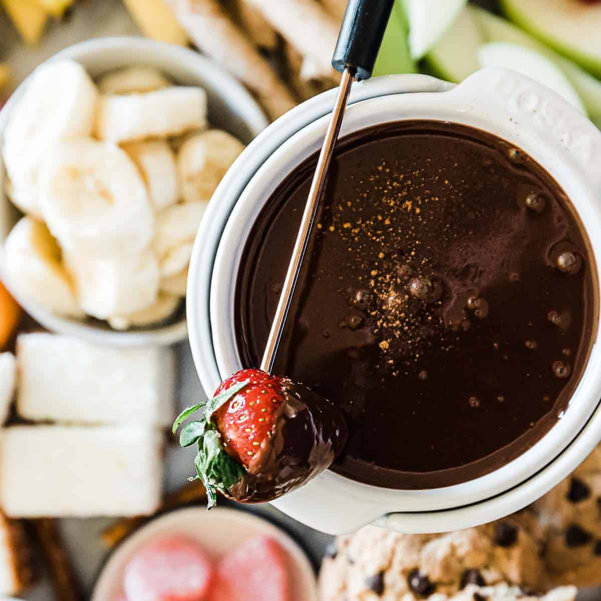 Fondue de chocolate em um pote de fondue branco rodeado de frutas.