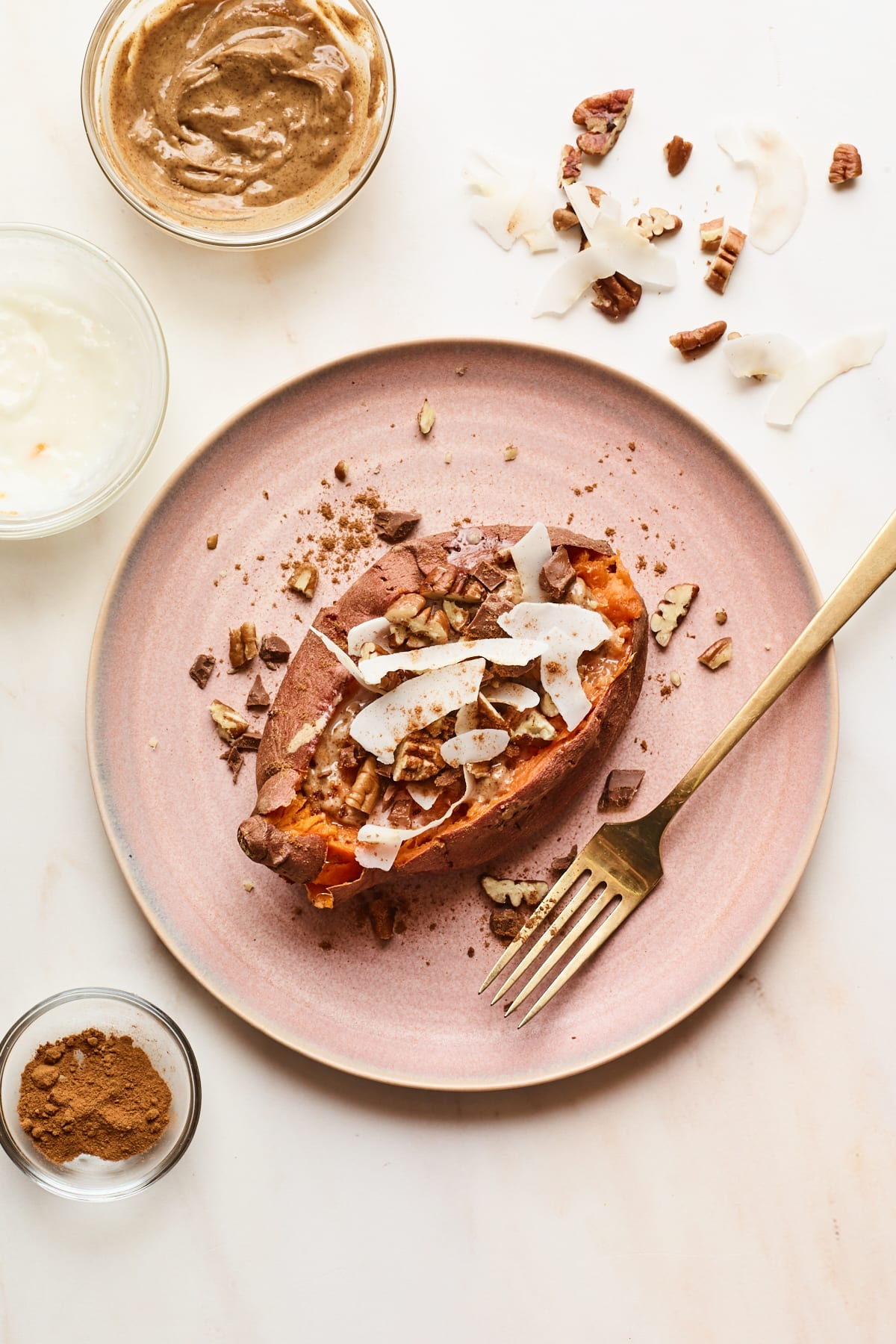 Um prato rosa com uma batata doce de sobremesa no topo. Os ingredientes de cobertura são polvilhados à volta do prato e do tampo da mesa. Um garfo dourado repousa sobre o prato.