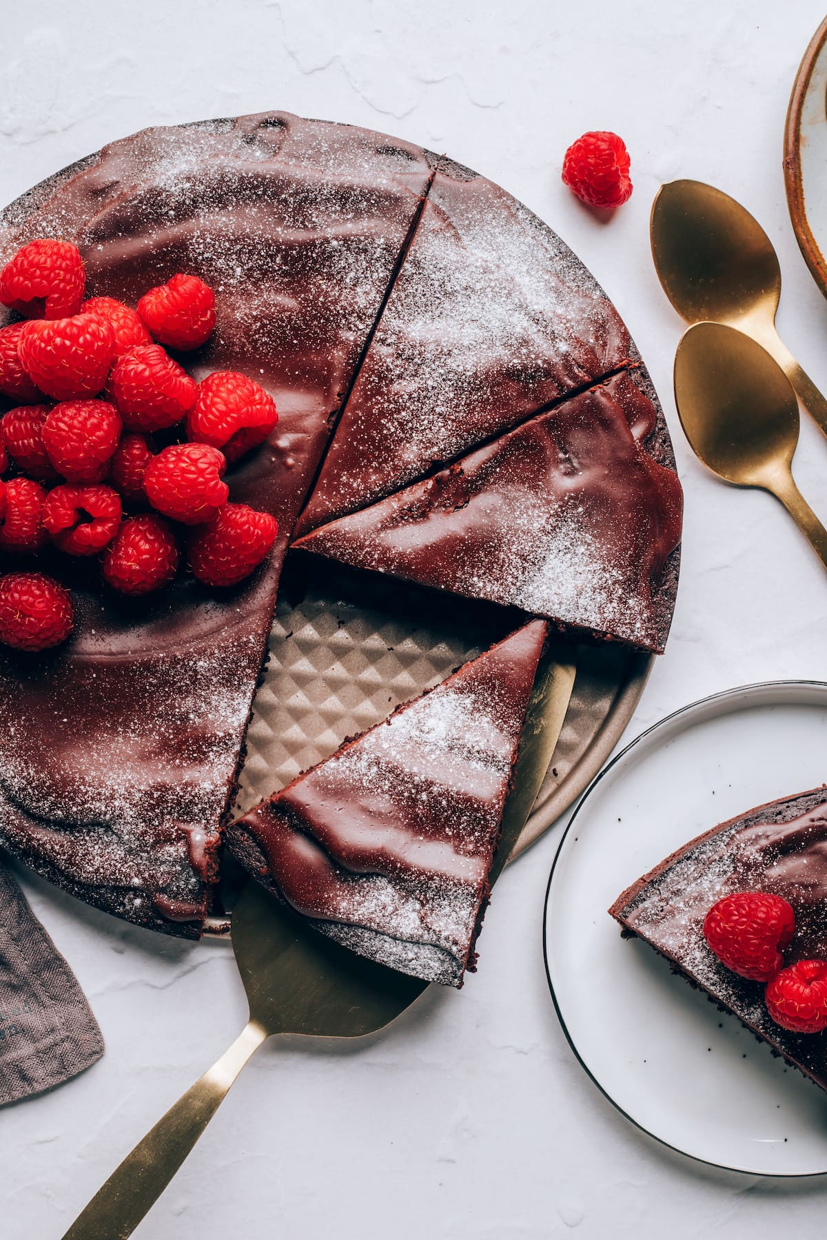 Uma vista aérea de um bolo de chocolate sem farinha, coberto com framboesas e açúcar em pó. Uma fatia de bolo está descansando sobre um utensílio para servir.