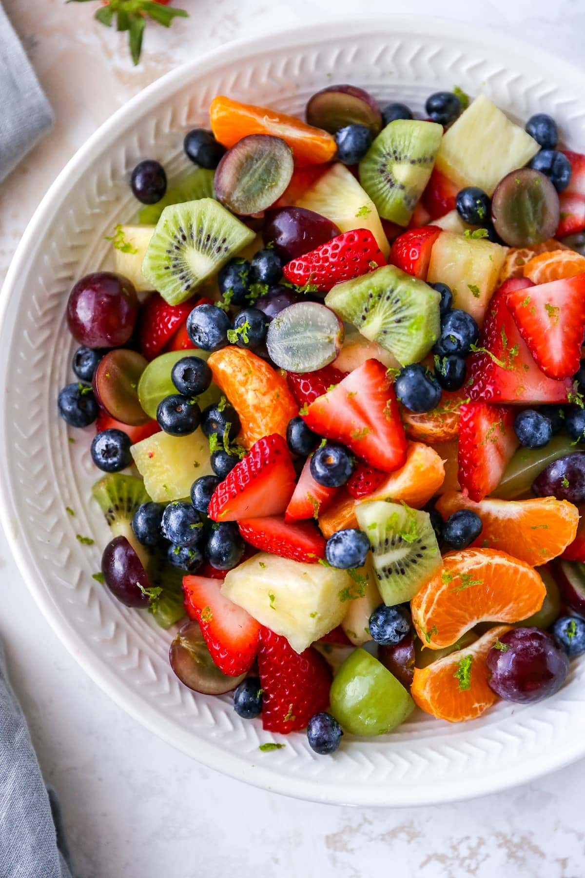 Tigela de salada de frutas com morangos, clementinas, kiwi, uvas, mirtilos e abacaxi. A salada de frutas é coberta com raspas de lima.