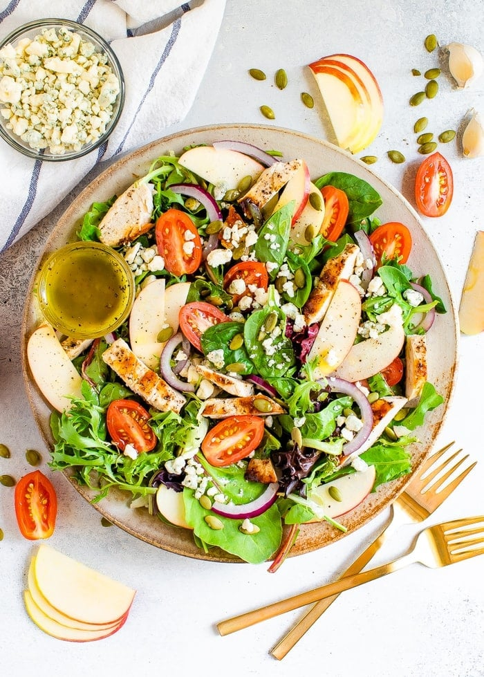 Prato com uma salada de frango com maçã fuji. Dois garfos, pepitas, maçãs, tomates e gorgonzola são polvilhados à volta do prato.