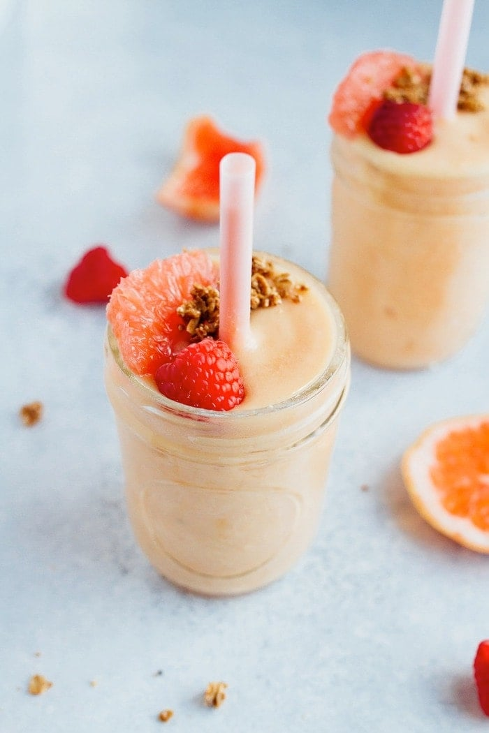 Dois smoothies de ananás de toranja com palhinhas cor-de-rosa, cobertos com granola, uma framboesa e uma fatia de toranja. Estão rodeados de framboesas e fatias de toranja no balcão.