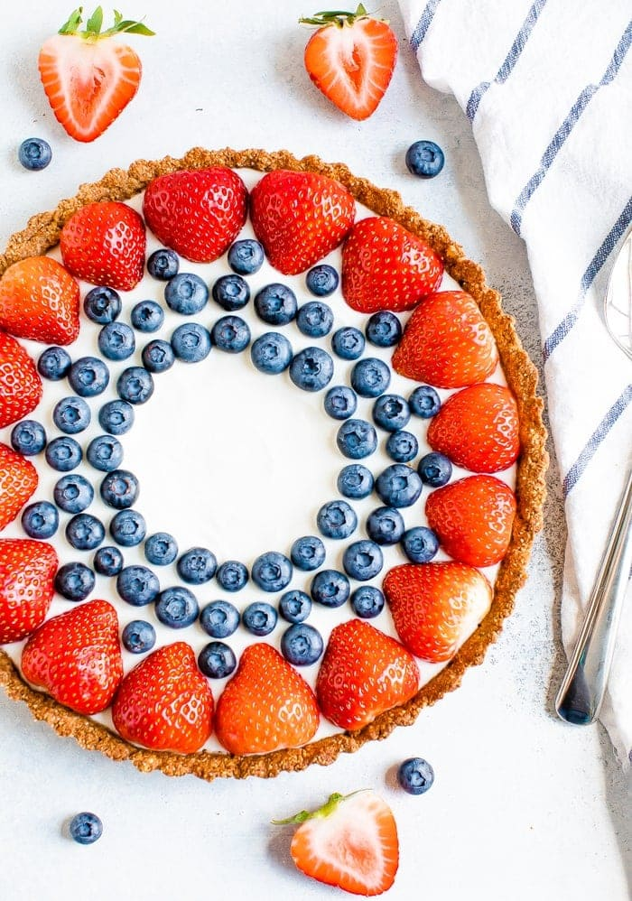 Torta grega de iogurte decorada com morangos e mirtilos. Uma toalha de prato às riscas, morangos fatiados e mirtilos rodeiam a tarte.