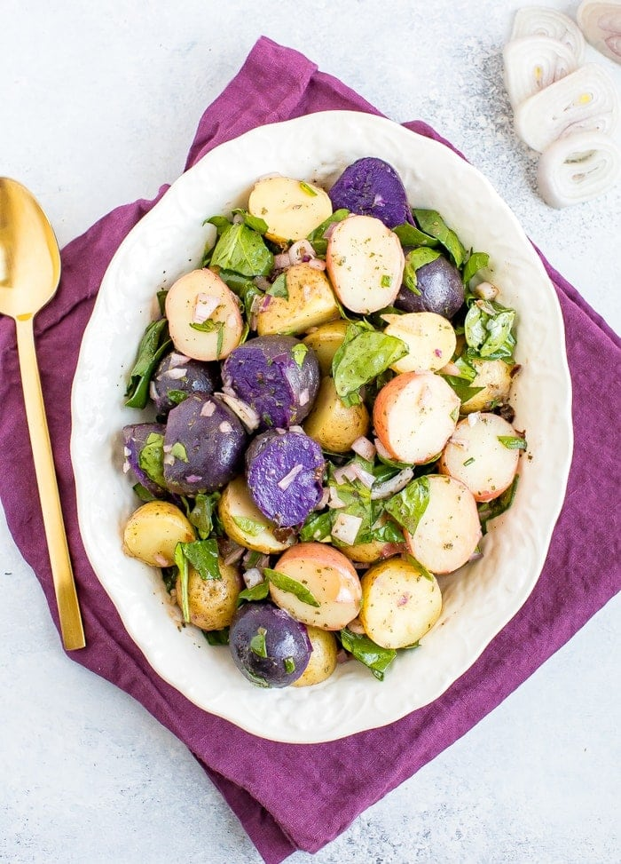 Salada de batata tricolor com chalotas e espinafres numa tigela branca com guardanapo púrpura e colher de ouro.