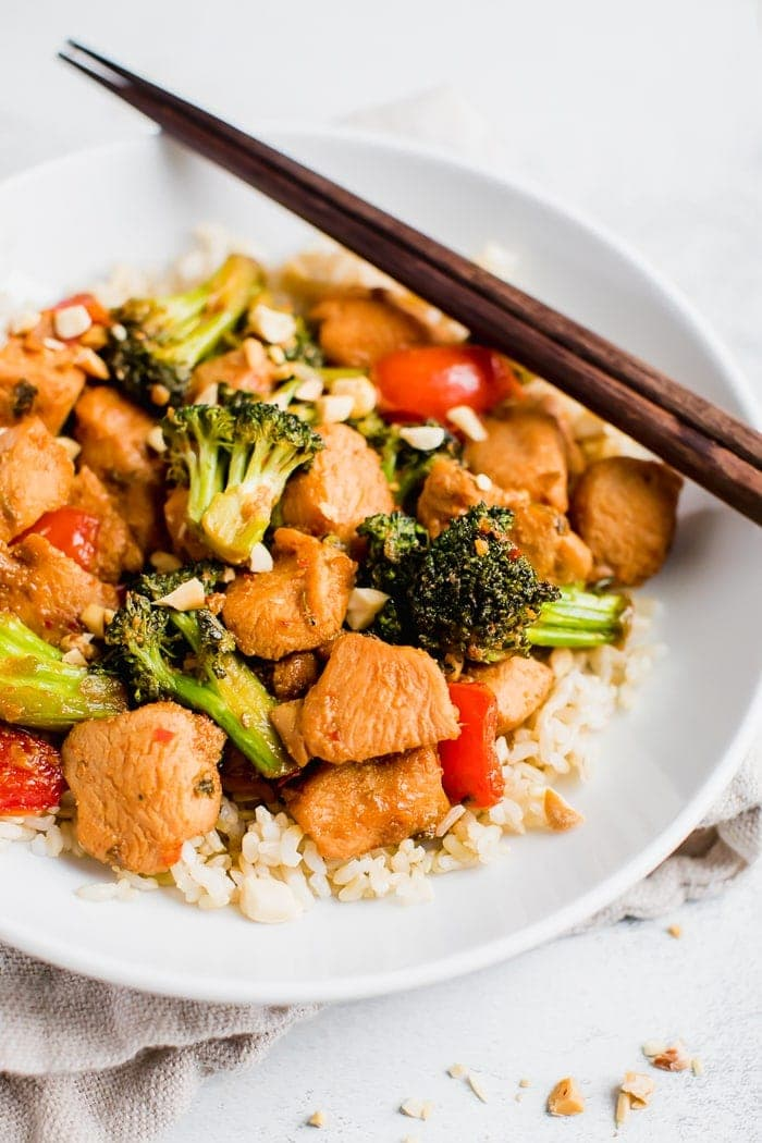 Frango Kung pao sobre arroz com legumes e amendoins em um prato com pauzinhos.