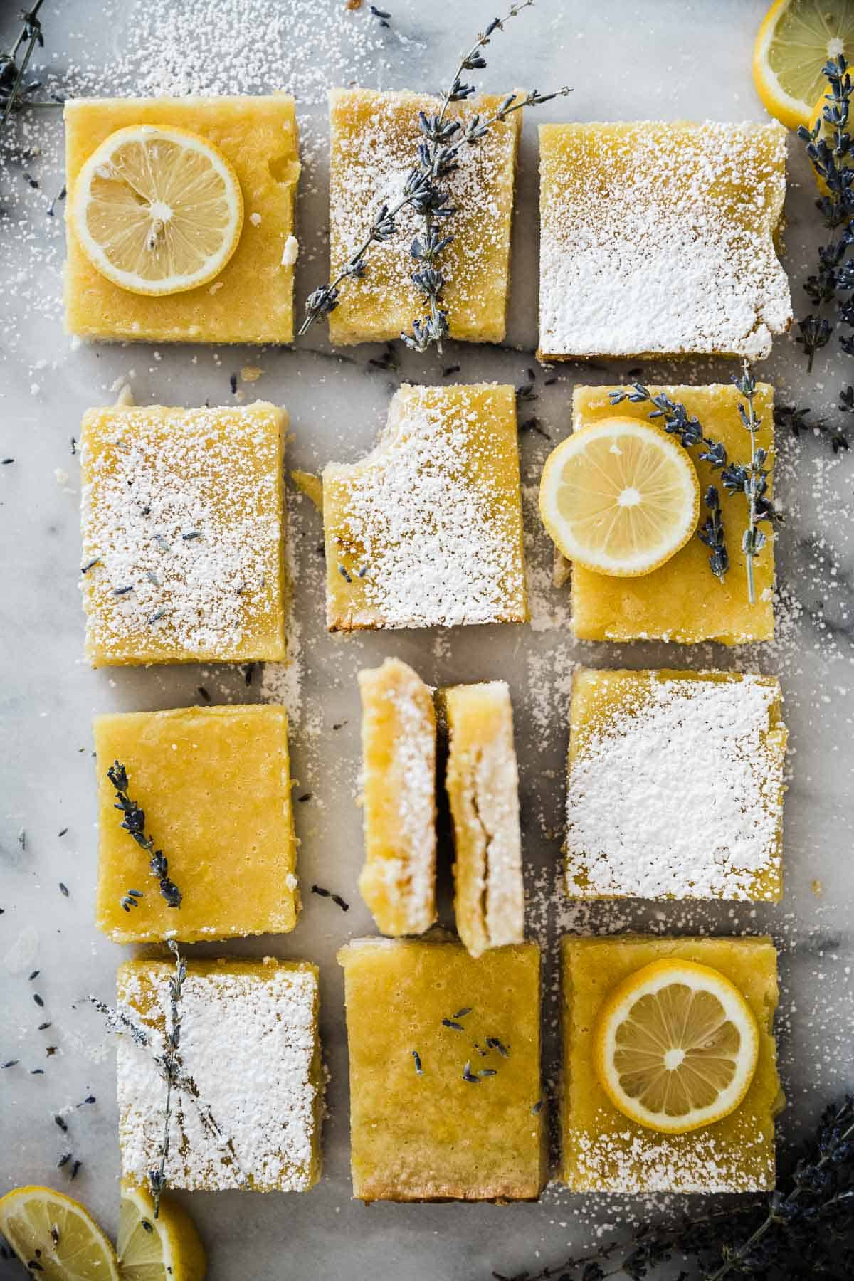 Barras de limão fáceis, cortadas em quadrados e colocadas sobre um balcão de mármore. Há algumas guarnecidas com fatias de limão, lavanda e açúcar em pó.