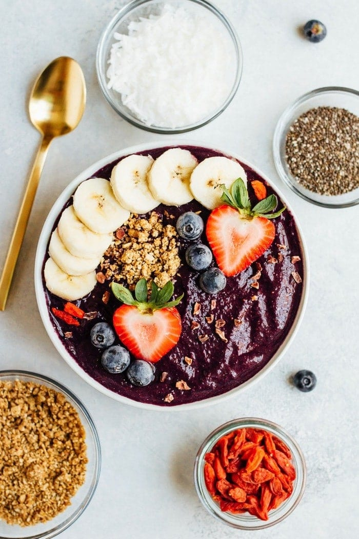 Taça de batido de bagas de Maqui coberta com banana, morangos, mirtilos e granola. Colher de ouro deitada ao lado da tigela juntamente com tigelas de bagas secas, granola, coco e chia.